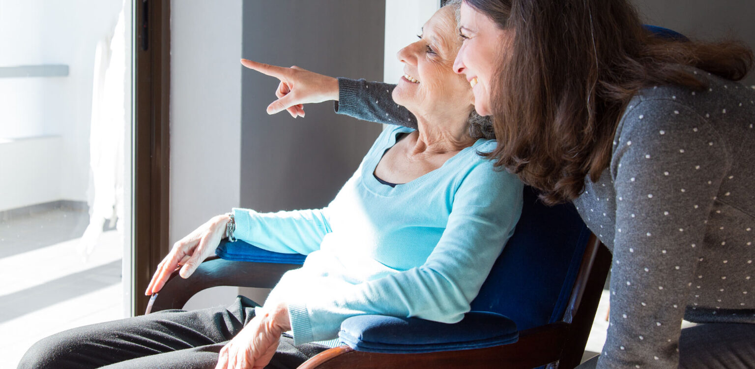Eine pflegebedürftige Seniorin genießt gemeinsam mit ihrer Tochter den Ausblick aus dem Fenster.