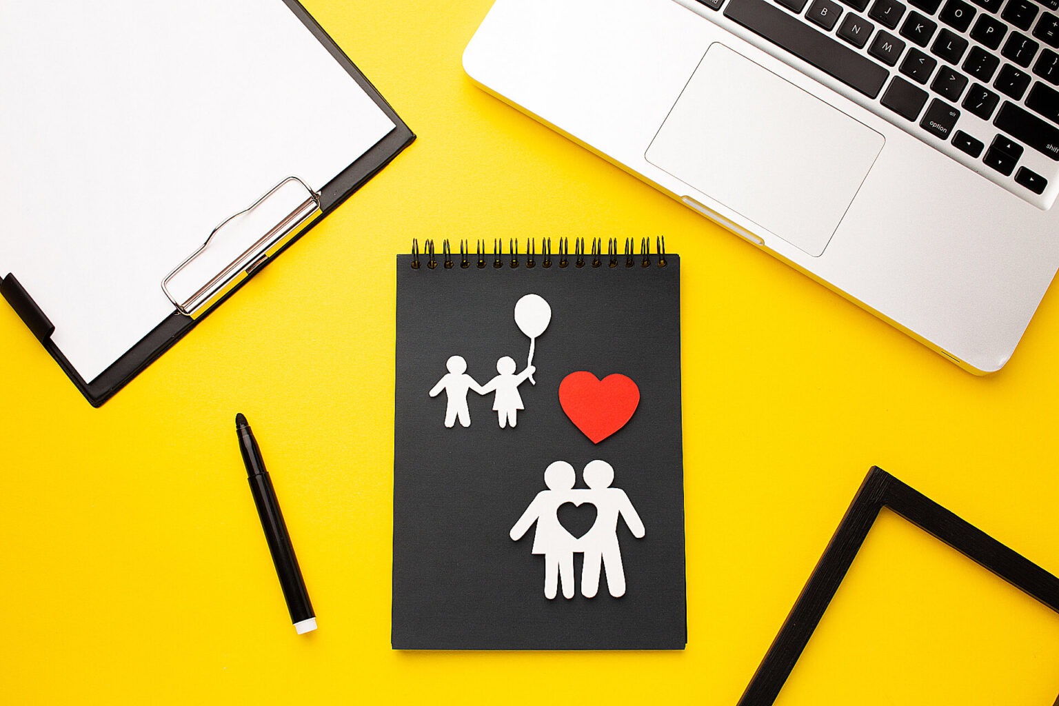 Blick auf einen Schreibtisch mit Klemmbrett und Notebook. In der Mitte ein Block mit ausgeschnittenen Figuren, die eine liebende Familie darstellen.